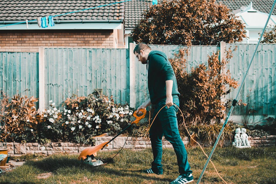 A man wearing glasses, a dark hoodie, and jeans is engaged in garden maintenance in a backyard, using a cordless electric grass trimmer to trim the lawn's edge. The lawn has lush green grass, with a s