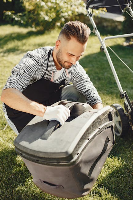 A man wearing glasses, a dark hoodie, and jeans is engaged in garden maintenance in a backyard, using a cordless electric grass trimmer to trim the lawn's edge. The lawn has lush green grass, with a s